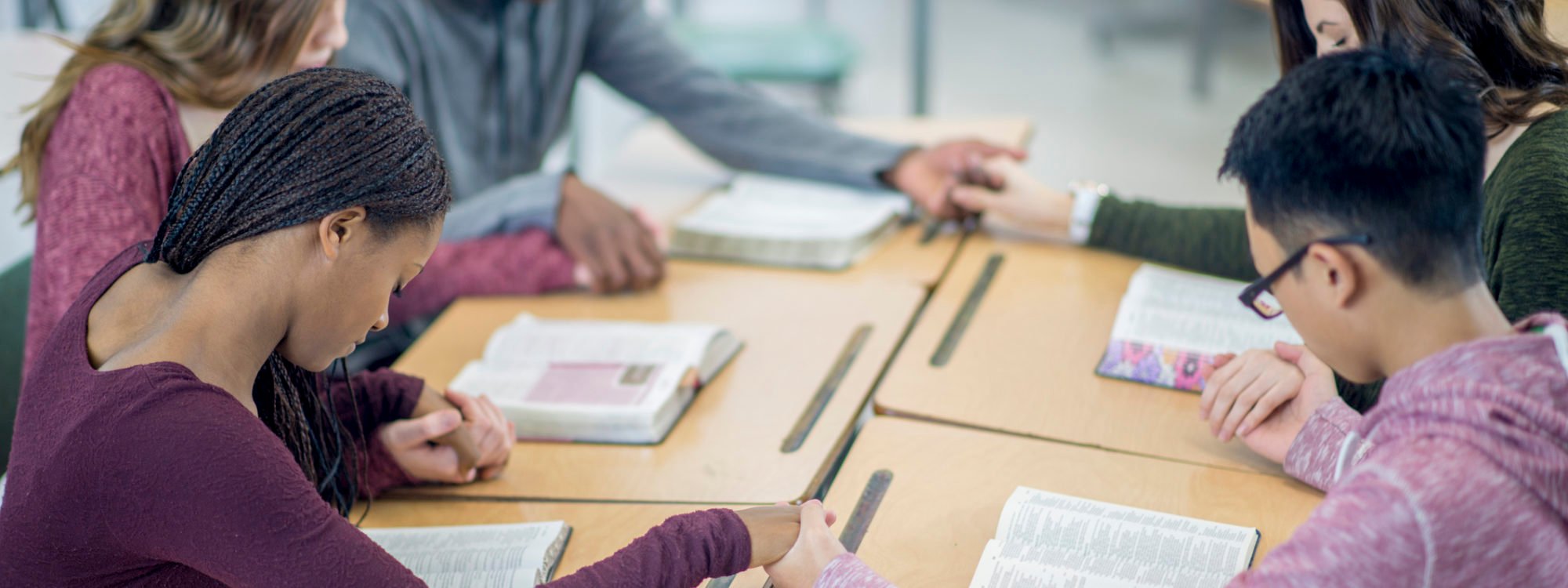 A multi-ethnic group of high school age students are sitting together at their desks and are holding hands while praying together.