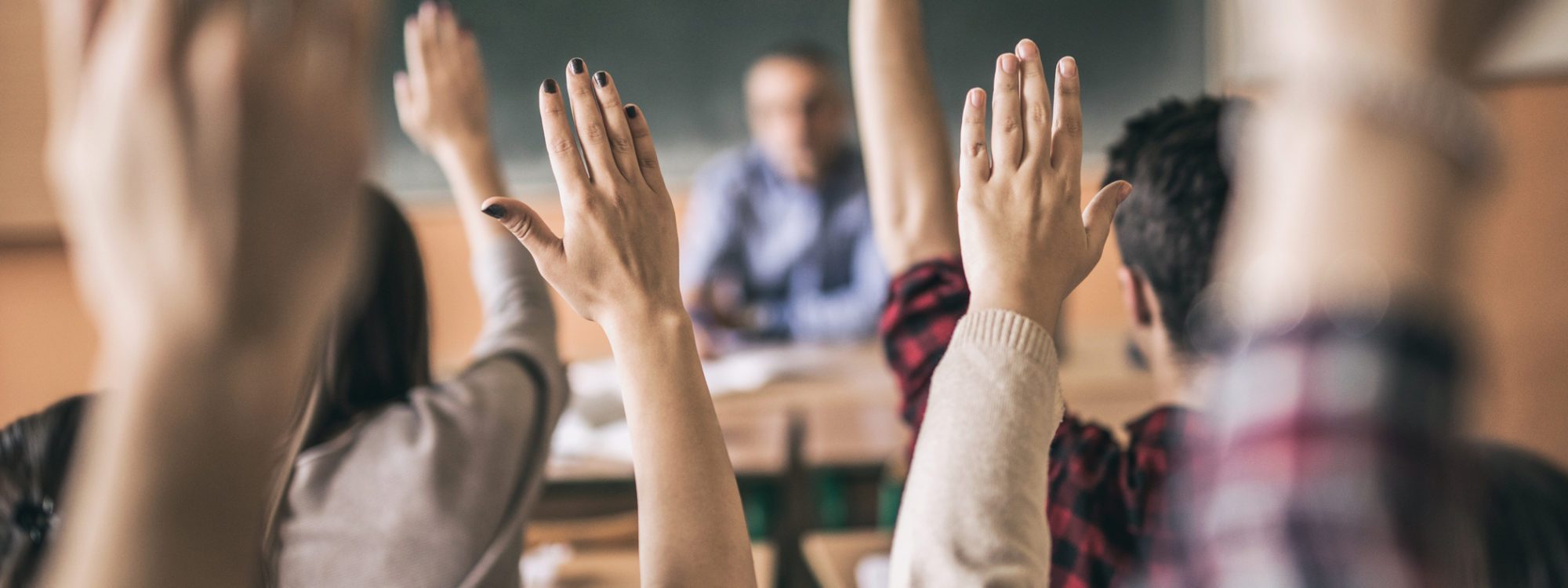 Rear view of group of students raising hands to answer teacher's question in the classroom. Focus is on hands in the middle.