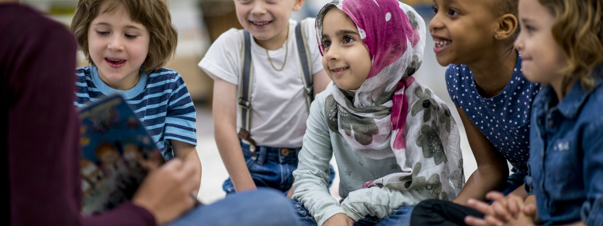 A multi-ethnic group of school children are indoors in a classroom. They are wearing casual clothing. They are sitting on the floor and eagerly listening to their teacher read a storybook.