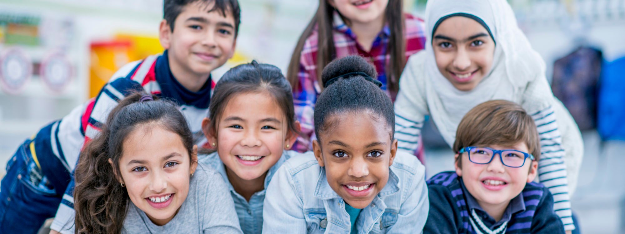 A diverse group of elementary school students are indoors in their classroom. They are lying on the floor and on each other. Everyone is smiling at the camera.