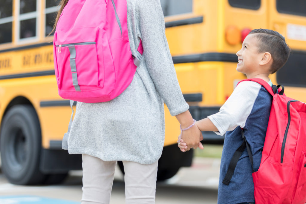 A kindergarten age little boy holds his unrecognizable older sister's hand and looks up at her with a smile as she guides him to his school bus on the first day of school.