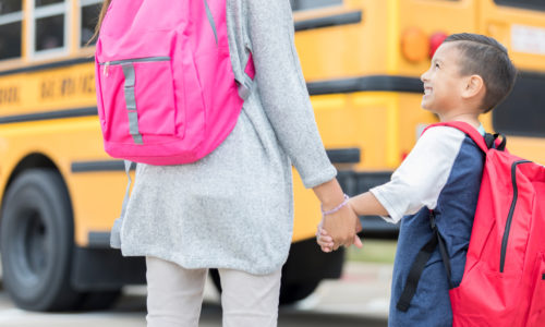 A kindergarten age little boy holds his unrecognizable older sister's hand and looks up at her with a smile as she guides him to his school bus on the first day of school.