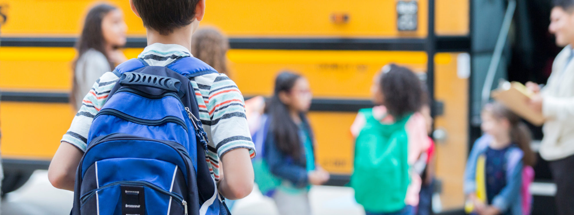 Rear view of elementary age boy waiting to get on school bus. His classmates are loading the bus in the background.