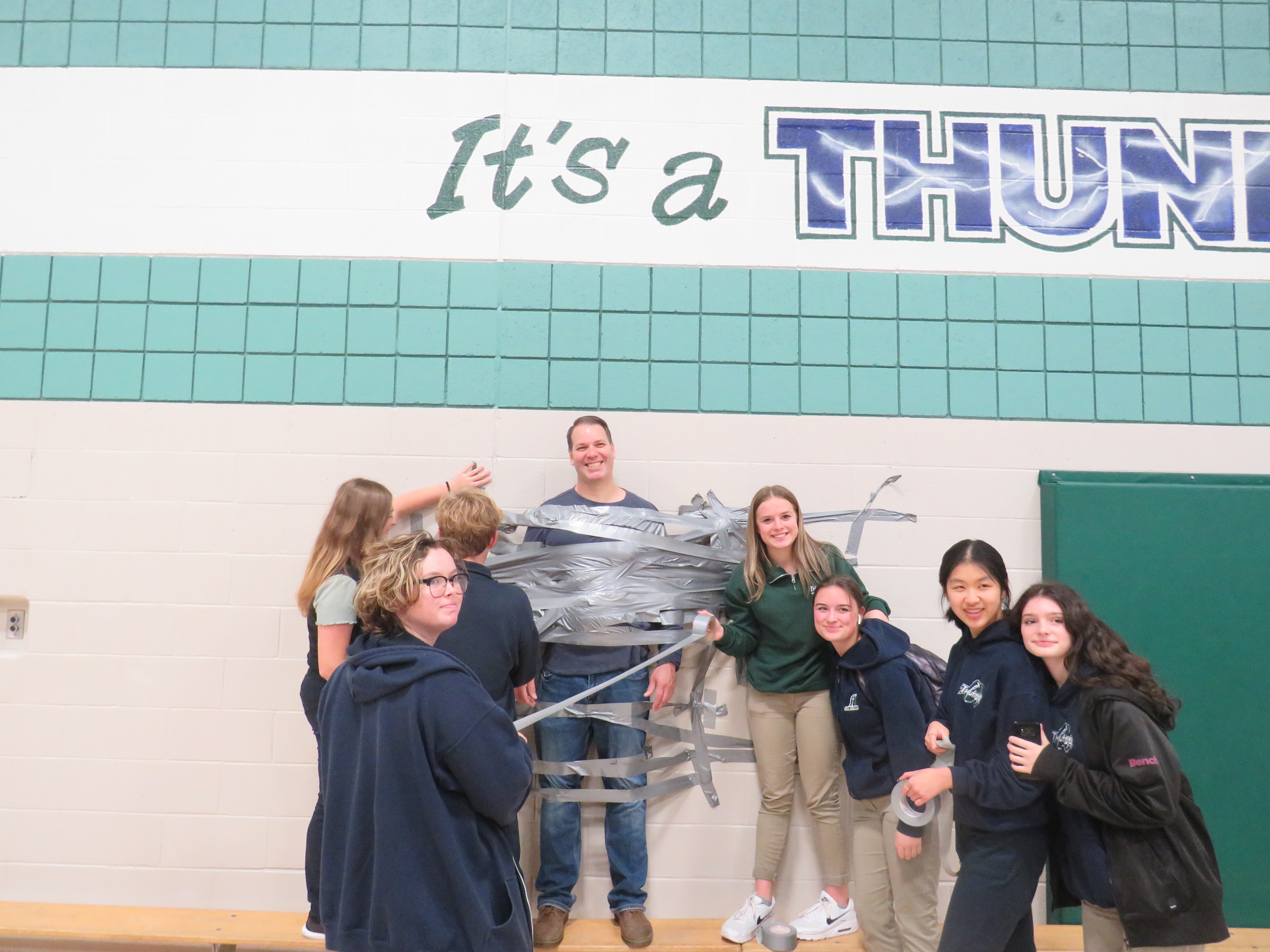 Students pose with teacher taped to wall for fundraising.