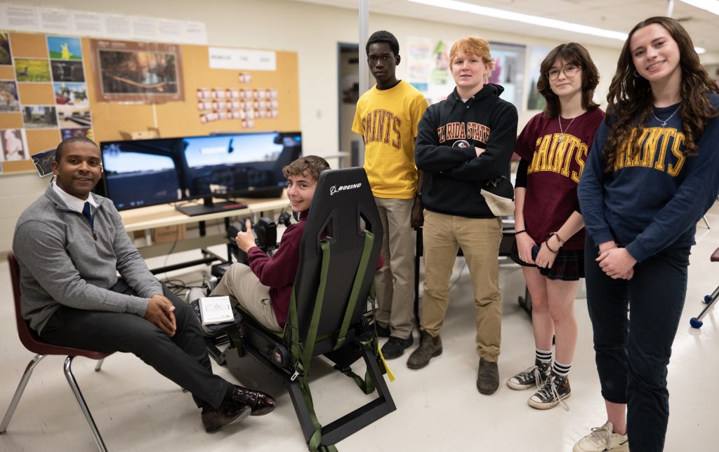 Students and an instructor gather around a flight simulator at a secondary school.