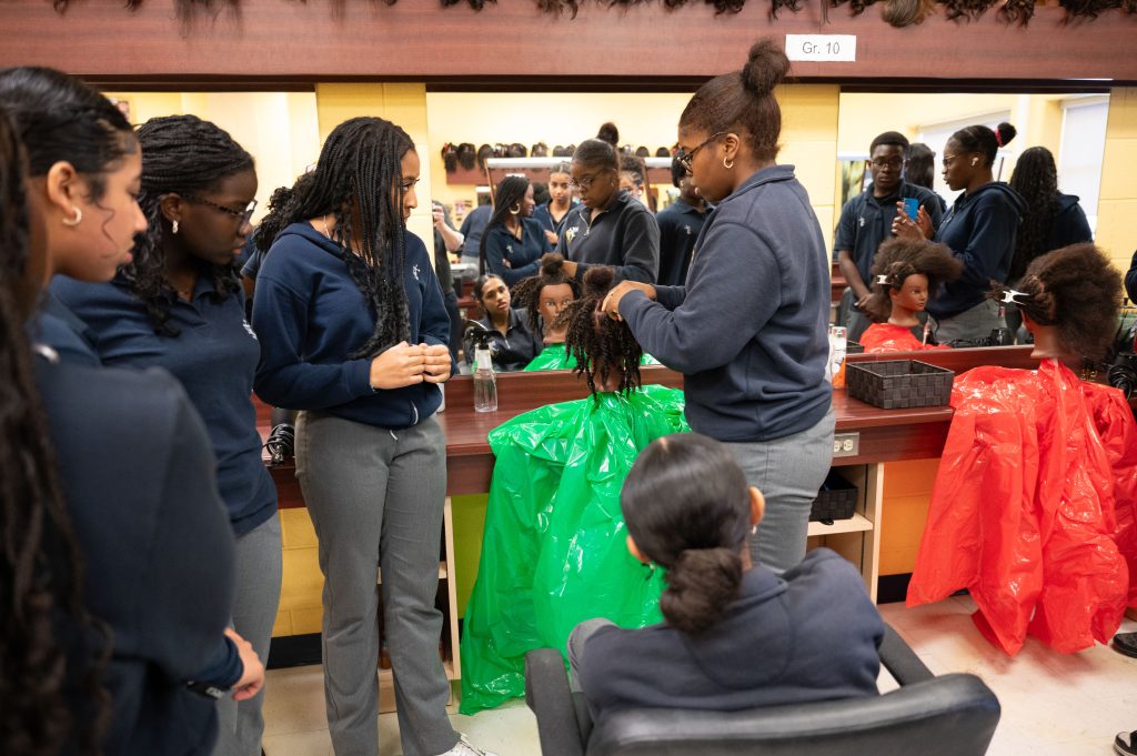 Students gather around a mannequin to learn how to braid hair.