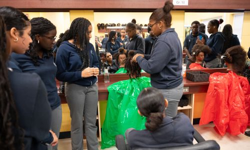 Students gather around a mannequin to learn how to braid hair.