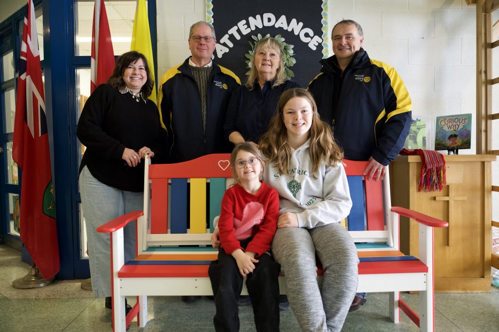 Group of adults and two students pose at a bench at a school.