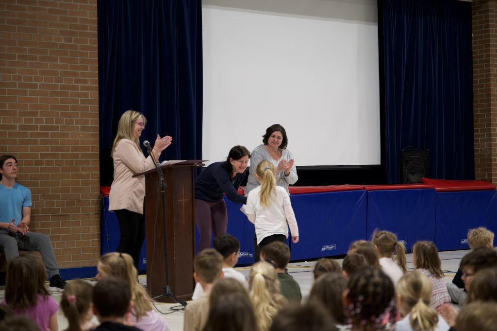 Student receives certificate from teacher while principal applauds and group of students looks on.