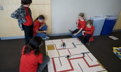 Four students and a coach look on as robot drives through a maze on the floor.