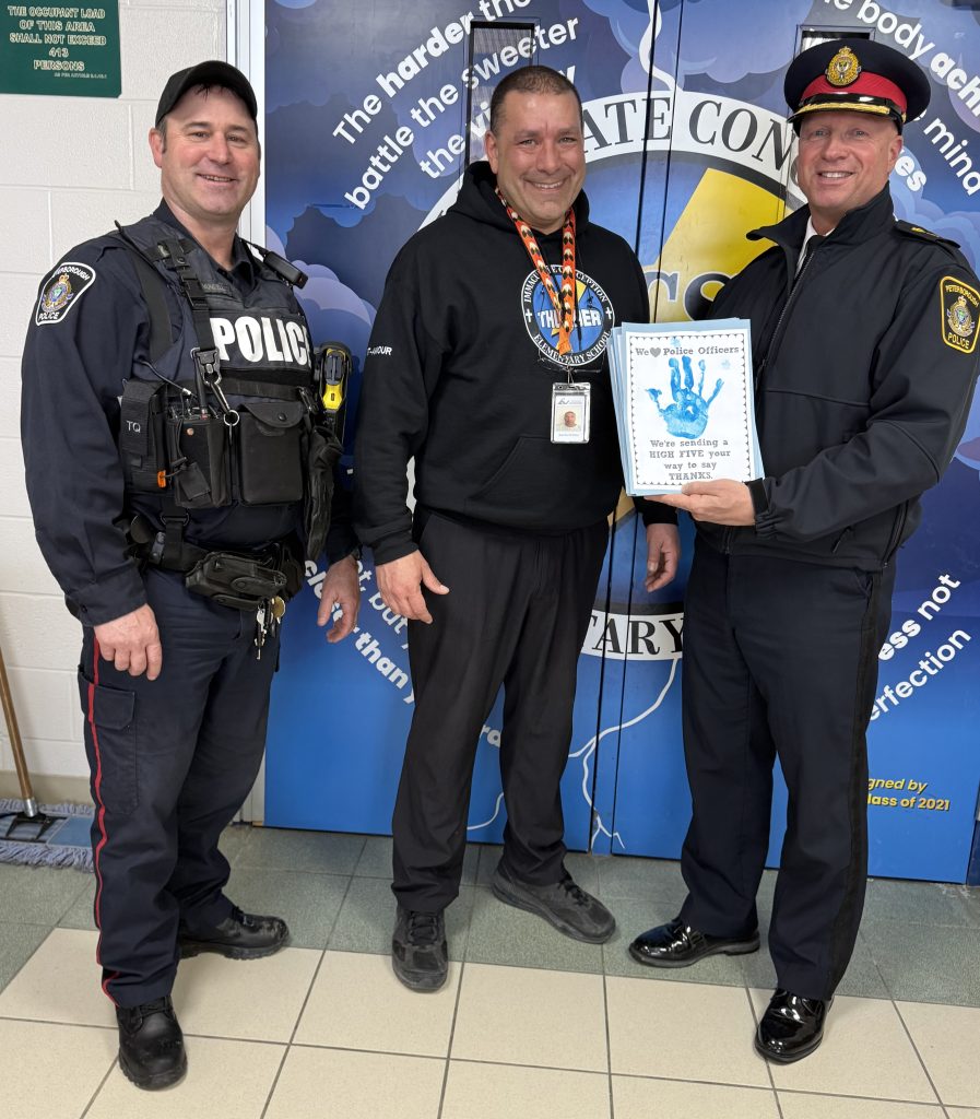 Two police officers stand with a principal at a school holding cards that were made for them by students.