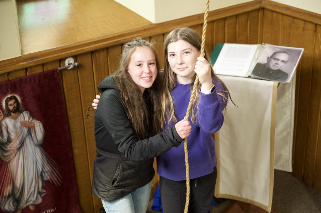 Two students pose for a photo inside of a church while holding the rope for the bell.