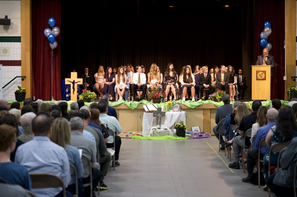 A group of students sitting on a stage at an awards ceremony in the gymnasium of a school.