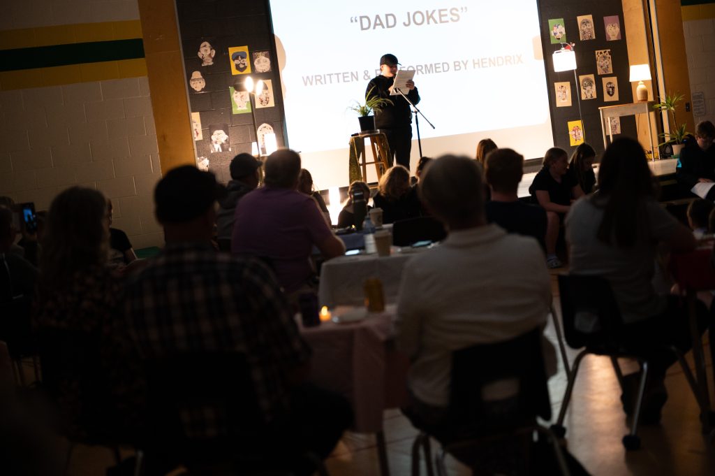 A student reads a poem in front of an audience at an assembly in a school gymnasium,