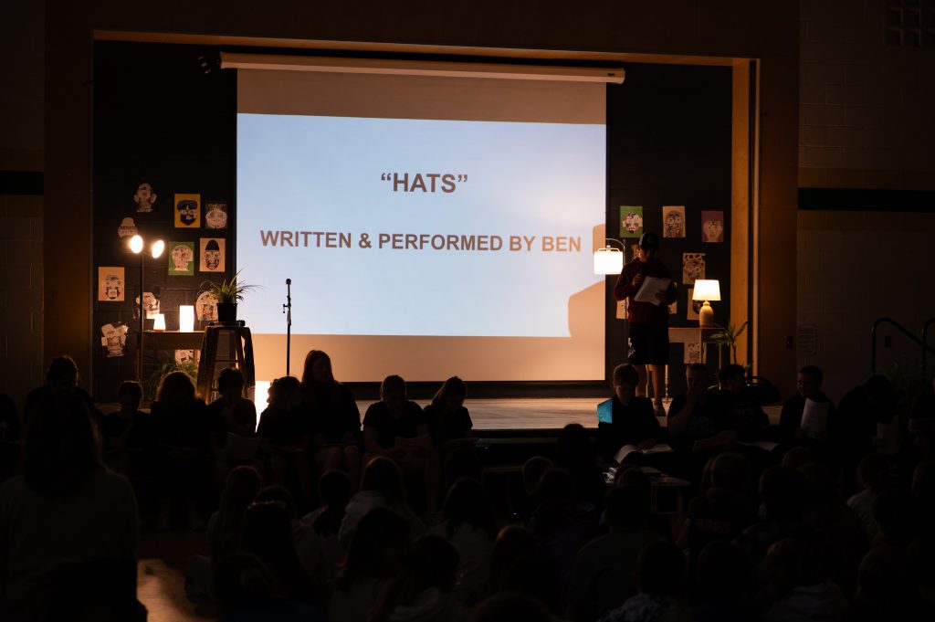 A student reads a poem in front of an audience at an assembly in a school gymnasium,