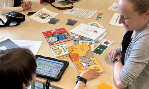 Child and adult read a book together while using a communication device.