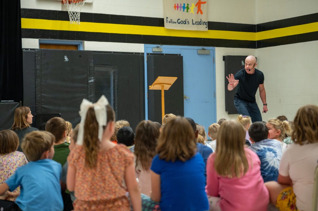Adult male stands in-front of a group of students at an assembly in a school gymnasium.