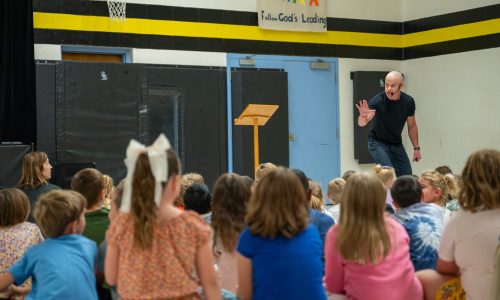Adult male stands in-front of a group of students at an assembly in a school gymnasium.