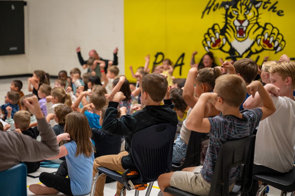 Group of students flex their muscles at an assembly in a school gymnasium.