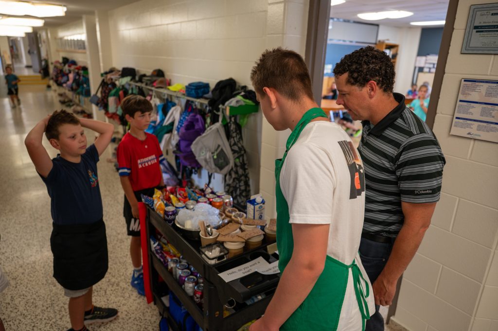 Three students serve treats from a coffee cart to an adult.
