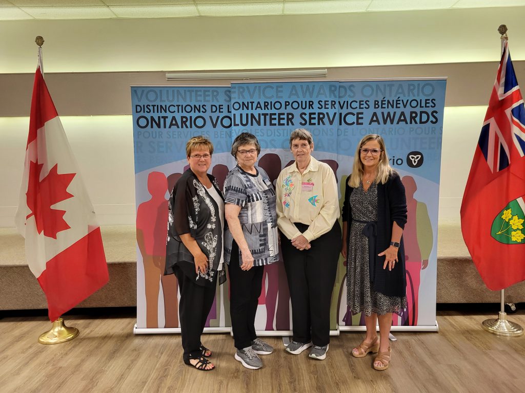 Four adult females stand in front of a background and flags of Canadad and Ontario at an awards ceremony.