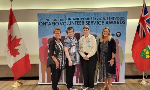 Four adult females stand in front of a background and flags of Canadad and Ontario at an awards ceremony.