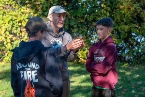 Two students look on as a man starts a fire using materials found in the forest,