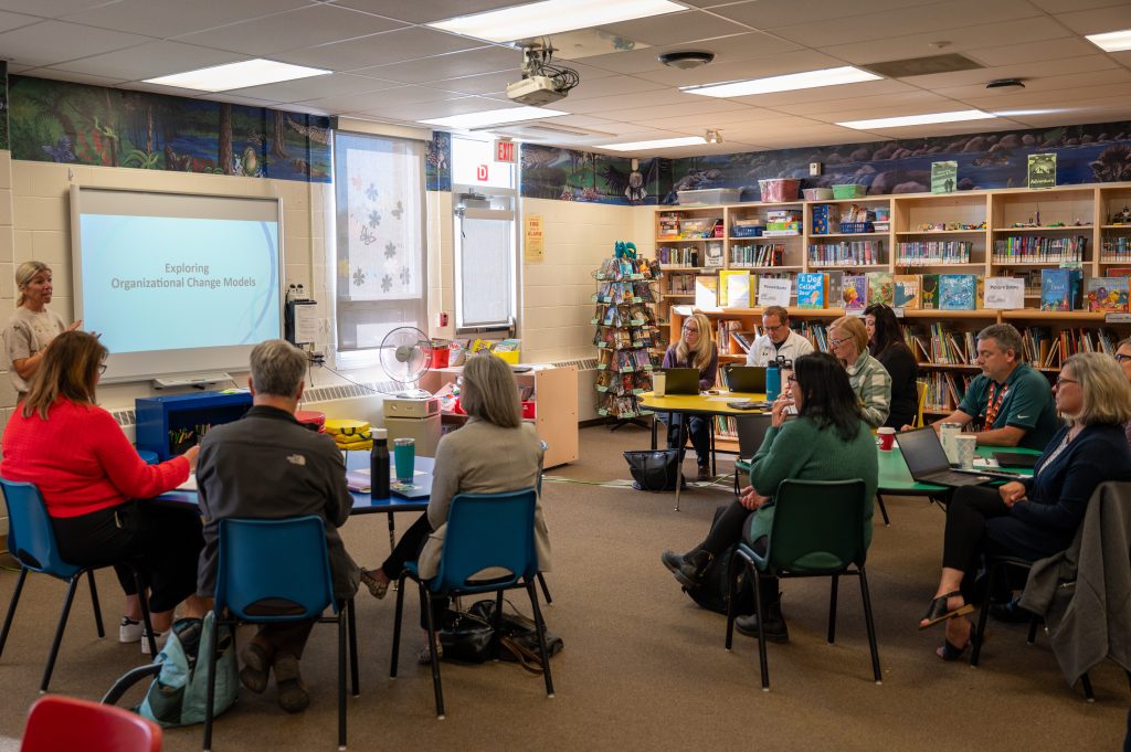 A group of adults sits in groups in a school library.