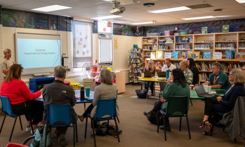 A group of adults sits in groups in a school library.