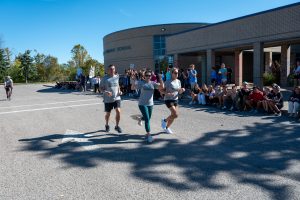 Runners jogging through a school parking lot while students cheer them on.
