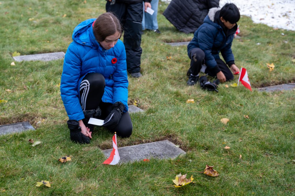 Two students kneel at the graves of fallen soldiers. Each student places a poppy and Canadian flag on the grave.