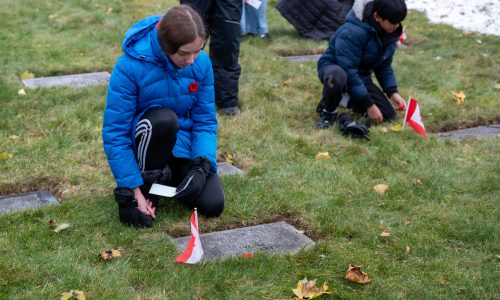Two students kneel at the graves of fallen soldiers. Each student places a poppy and Canadian flag on the grave.