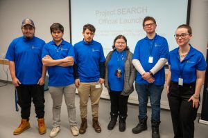 Six students post together in front of a projector screen.