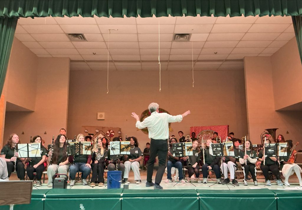 A large group of students on wind instruments plays on a stage at a school concert.