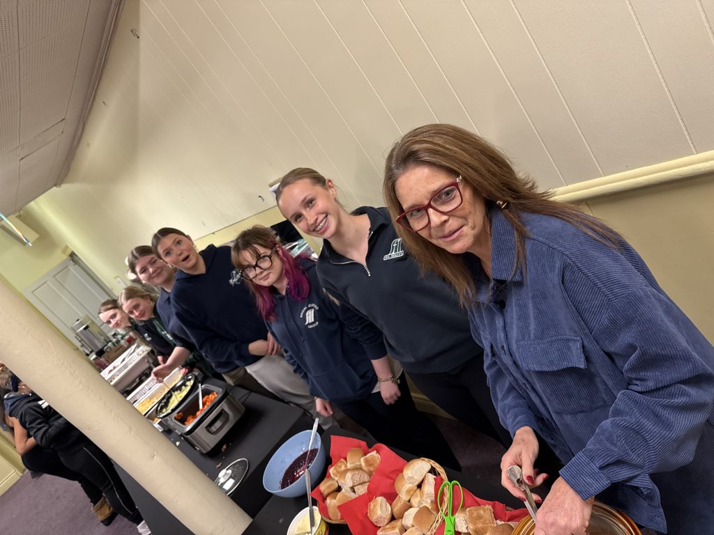 Students and staff members line a table where they prepare food for a community gathering.