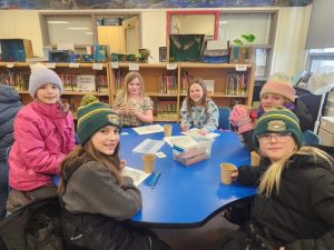 Students, wearing winter clothing, sit around a table inside a classroom with hot chocolate.