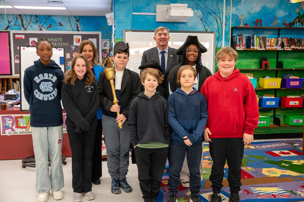 Students pose with the Speaker of the Ontario Legislative Assembly and the local Member of Provincial Parliament during a presentation at a school.