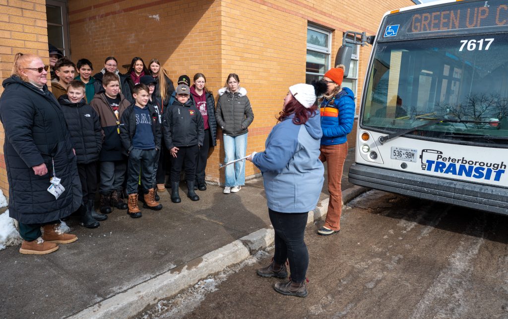 Students gather outside of a school to learn about transit while a city bus is parked nearby.