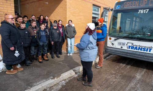 Students gather outside of a school to learn about transit while a city bus is parked nearby.