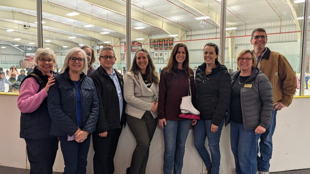 A group of adults stand in front of the glass of the ice surface in a hockey arena.
