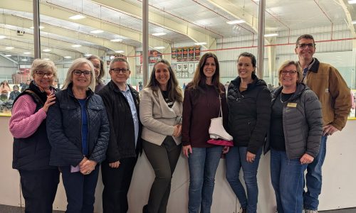 A group of adults stand in front of the glass of the ice surface in a hockey arena.