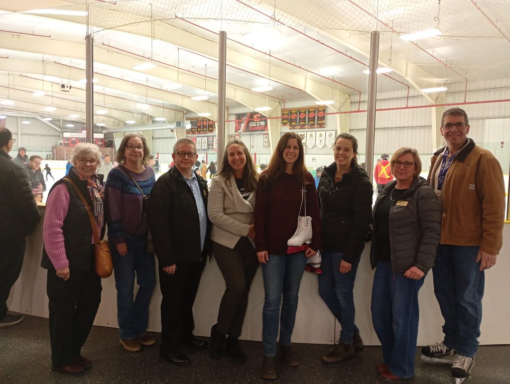 A group of adults stand in front of the glass of the ice surface in a hockey arena.