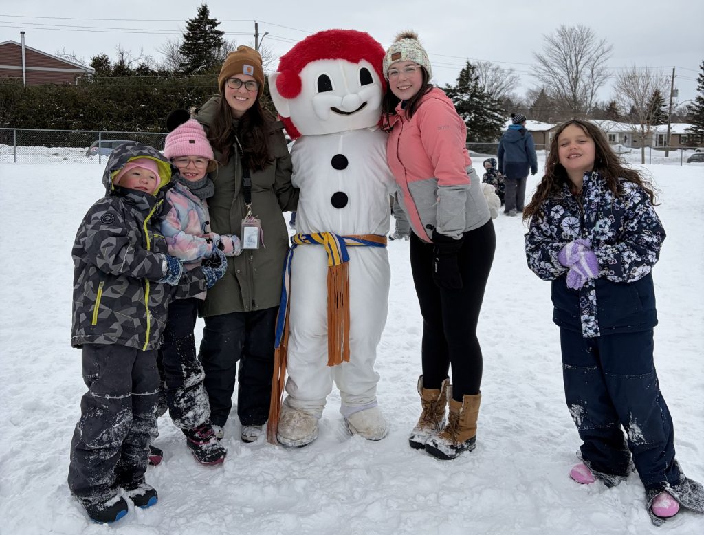 Two adults and three children stand with a snowman mascot at a school winter carnival.