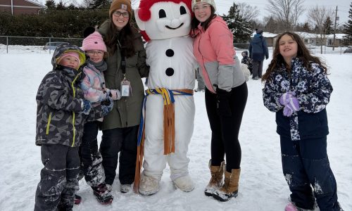 Two adults and three children stand with a snowman mascot at a school winter carnival.