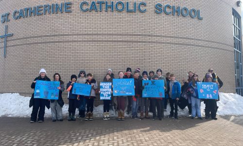 Students holding blue signs stand outside of a school on a winter day.