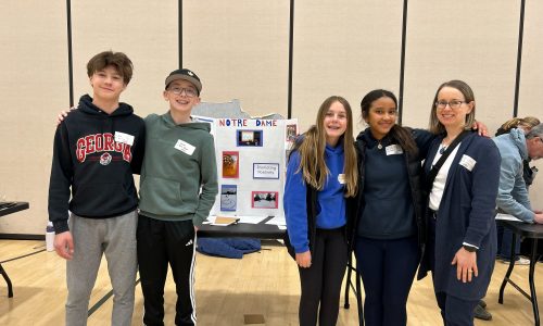 A group of four students and their teacher stand in front of a project board at a Student Summit.