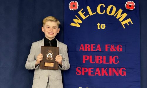 A male elementary school studnet stands in front of a sign at a Public Speaking competition while holding an award.
