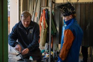A student wearing welding gear speaks with his teacher at a skilled trades event.