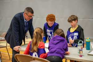 An educator looks on as four students solve math problems at a competition in a school gym.
