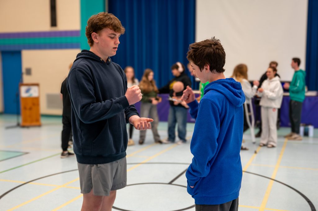 Two male students play a game of Rock, Paper, Scissors while standing in a school gymnasium.